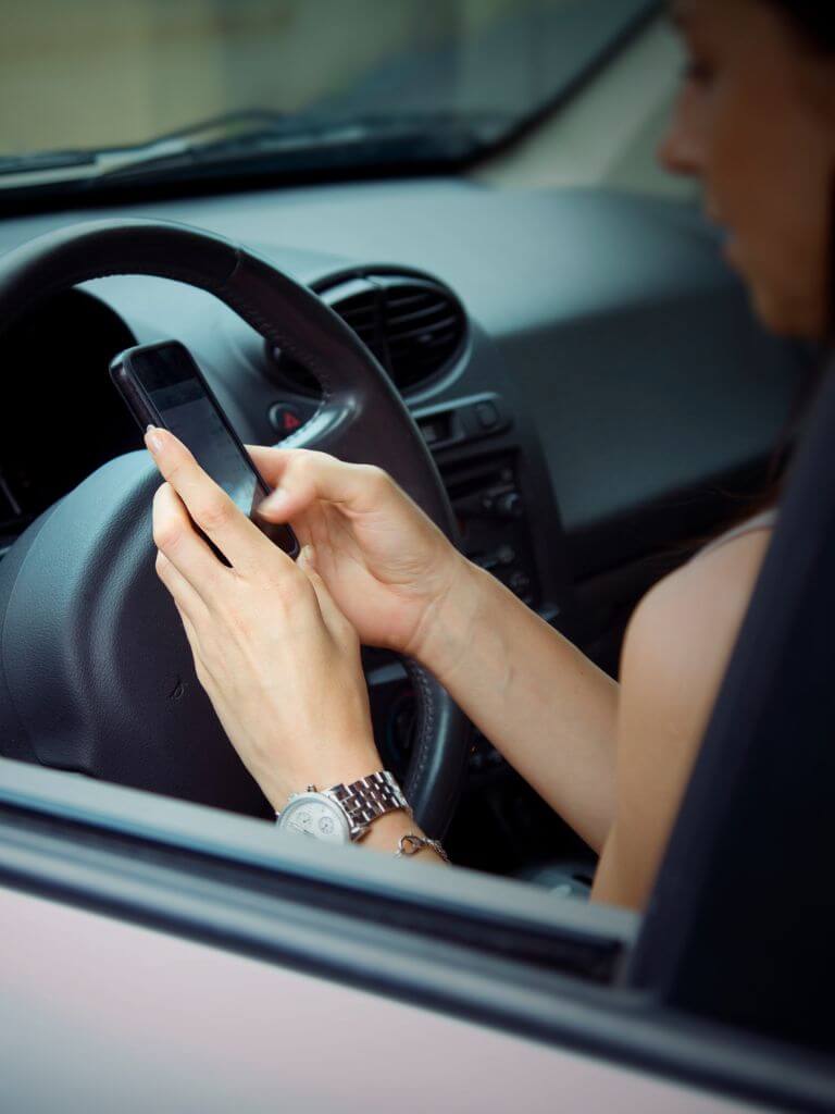 Person sitting in a car driver's seat, holding a smartphone with both hands, wearing a wristwatch.