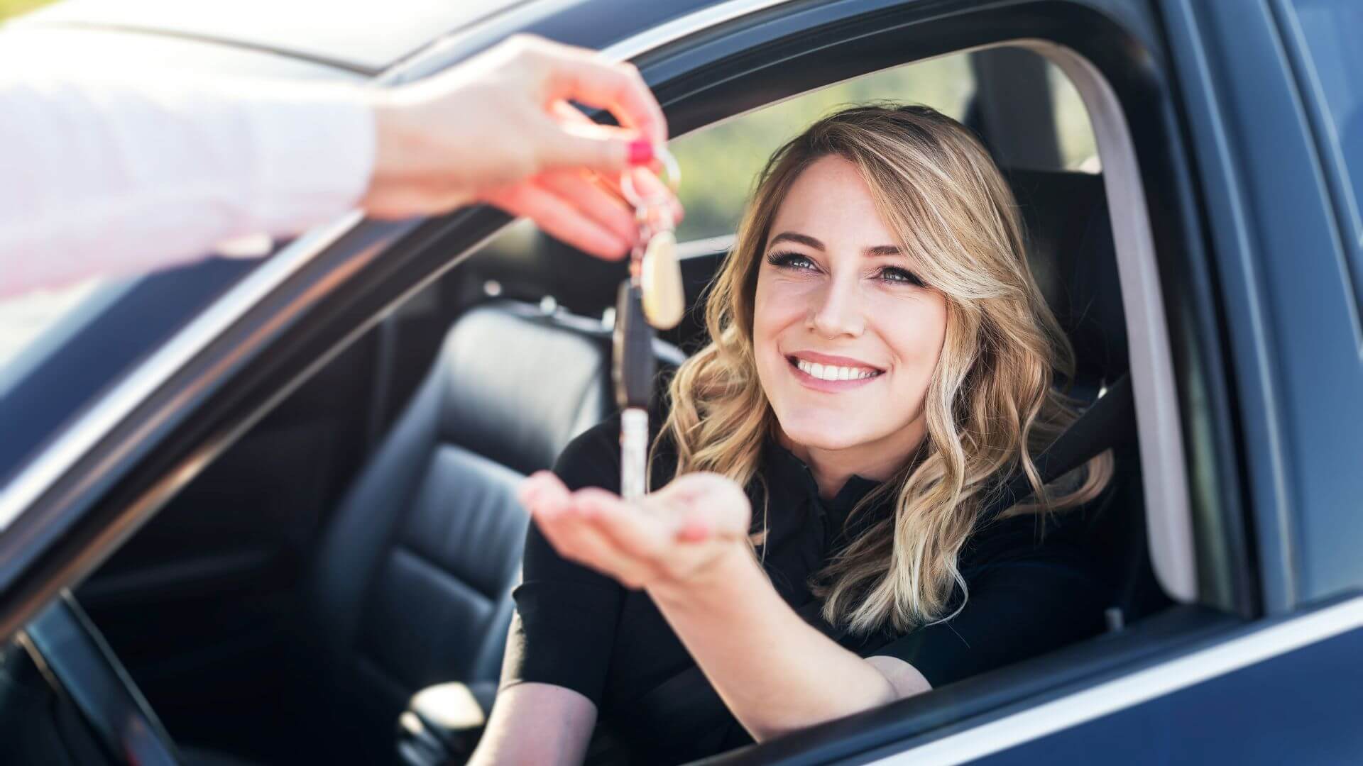A smiling woman sits in a car, reaching out to receive keys from a person's hand through the window, grateful for the efficient towing services that made her journey smooth.