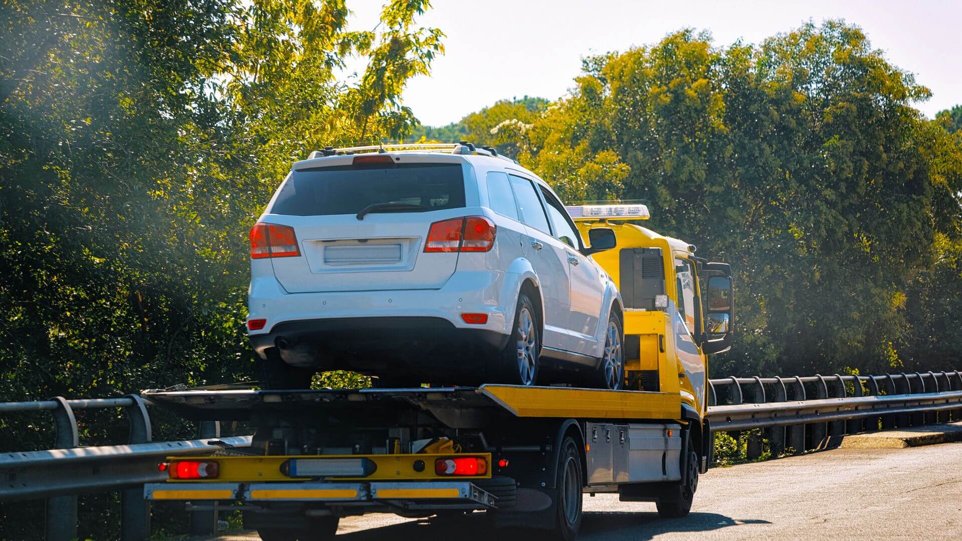 A tow truck carrying a white SUV rolls steadily down the rural road, flanked by lush trees, evoking a sense of home nestled amid tranquility.