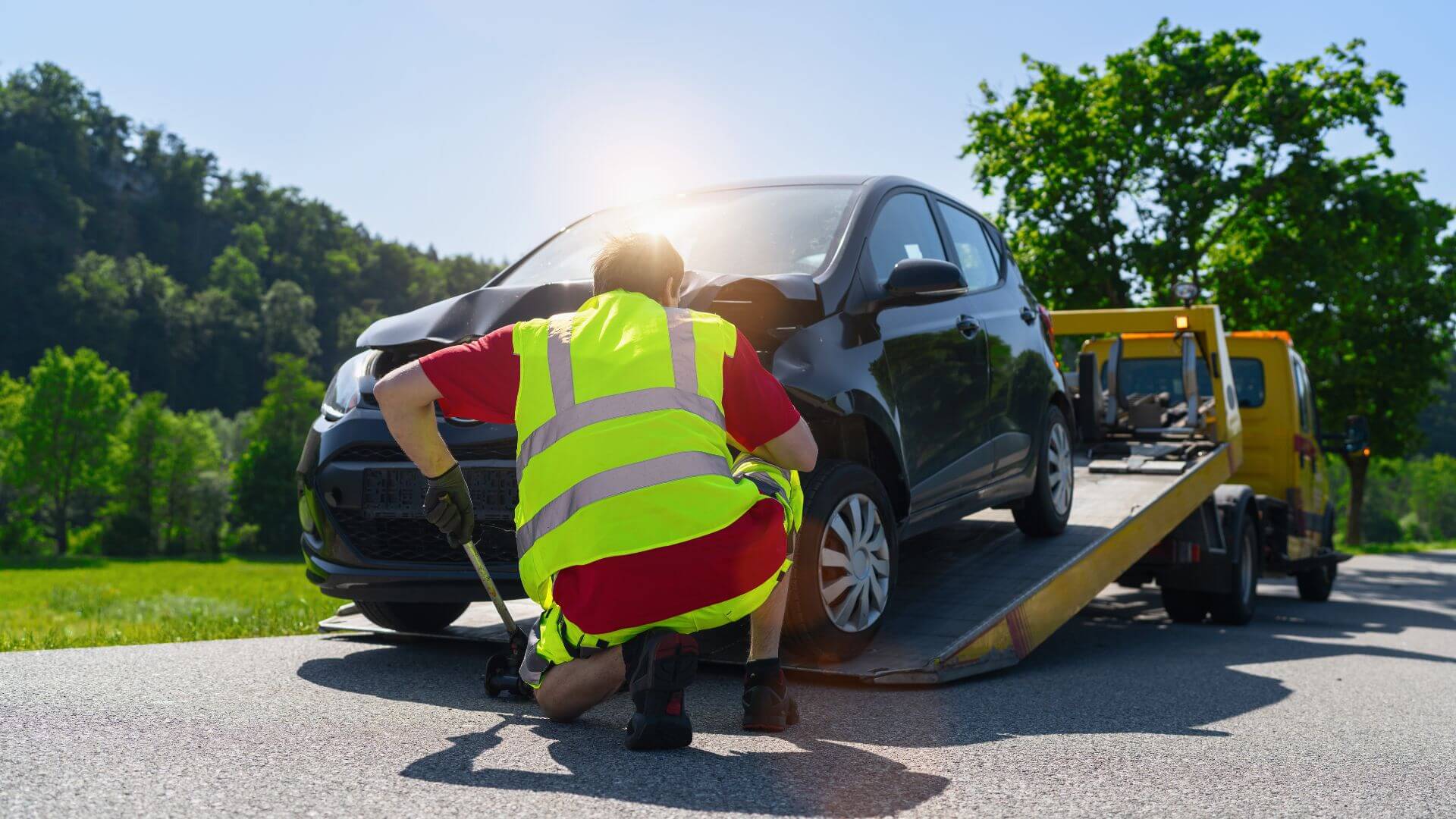 A worker in a high-visibility vest secures a black car onto a tow truck on a sunny rural road, showcasing the efficiency and reliability of professional towing services.