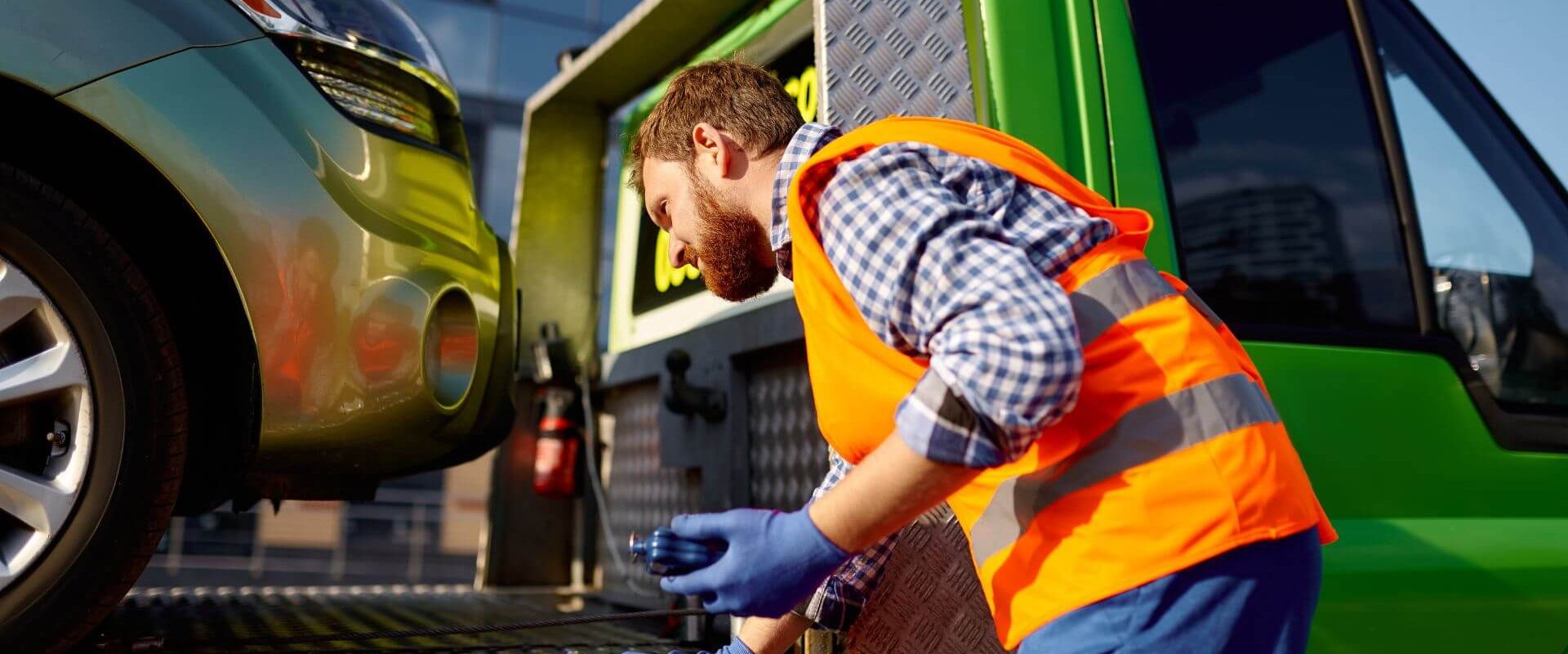 A man in a reflective vest and gloves carefully inspects a green car on a tow truck, ensuring its safe journey home. A man in a reflective vest and gloves carefully inspects a green car on a tow truck, ensuring its safe journey home.